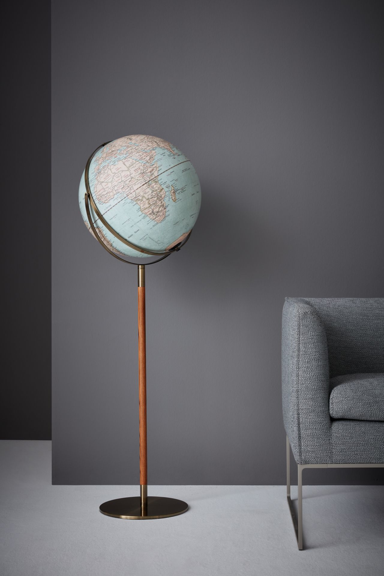 Standglobus des Herstellers Emform mit elegantem Holzgestell auf einem Metall Tellerfuß. Der Globus steht vor einer grauen Wand und neben einem Sofa.