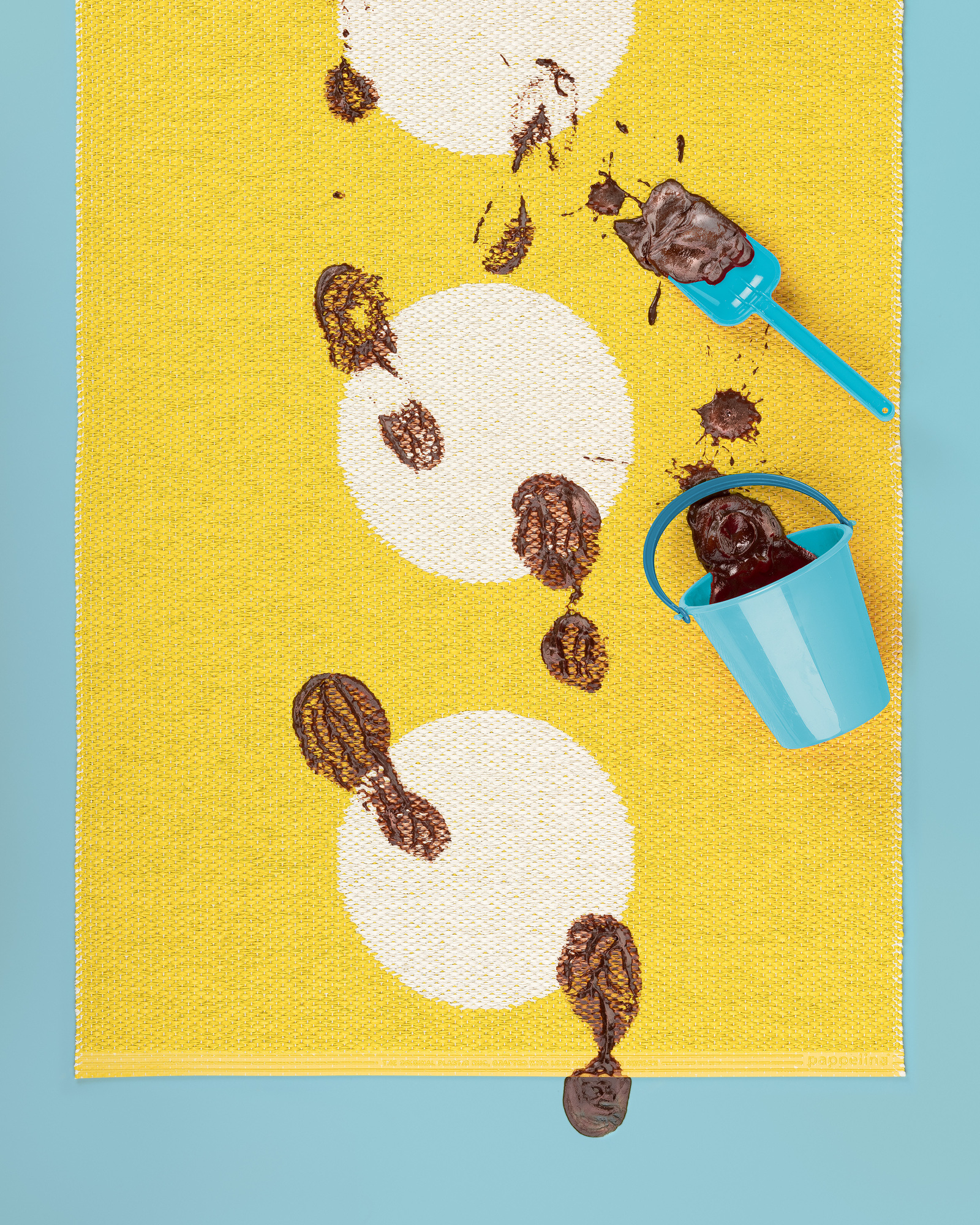 Vera Kunststoffteppich Pappelina in Gelb mit weißen Kreisen, dekoriert mit Schokoladenfußabdrücken, Eimer und Schaufel.