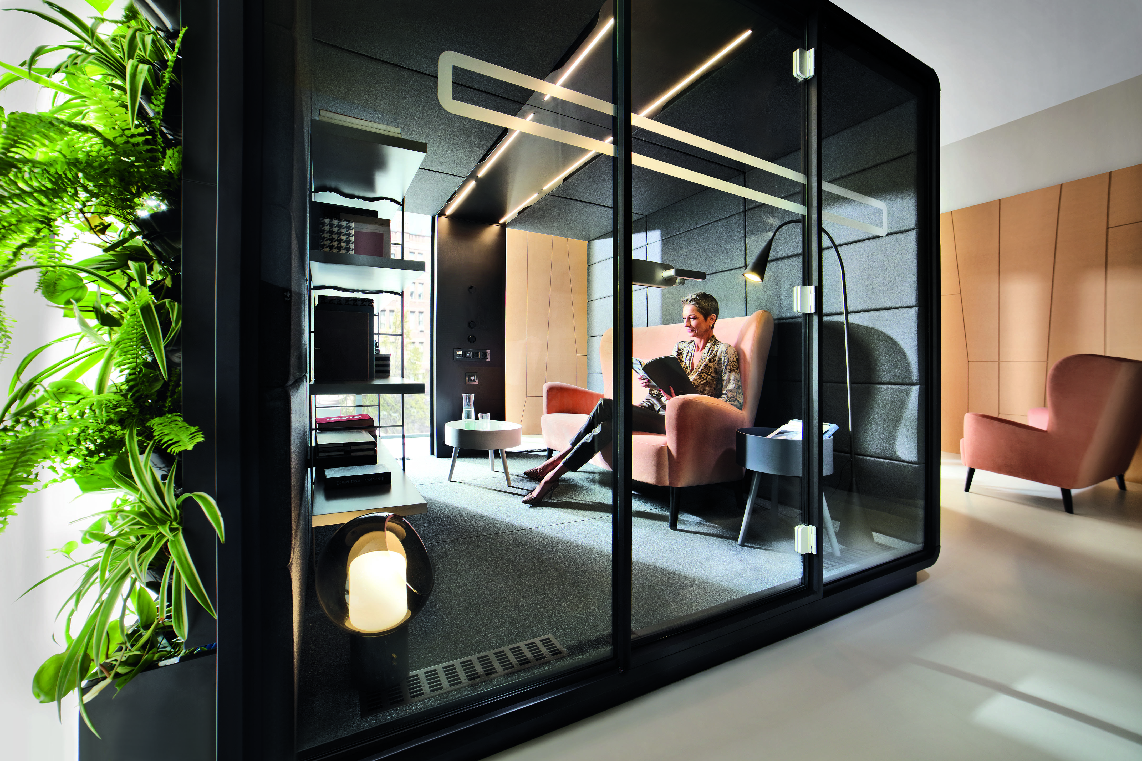 HushMeet L Meetingbox: Frau liest in moderner Telefonzelle/Besprechungsraum mit Glaswänden und Pflanzenwand.